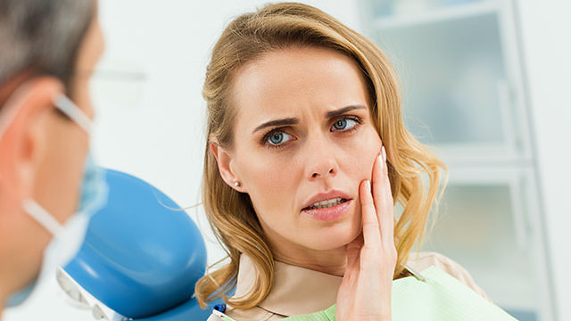 A concerned woman with blonde hair is sitting in a dental chair, touching her cheek with a pained expression. She appears to be discussing her discomfort with a dentist, who is partially visible in the foreground wearing a mask. The setting is a dental clinic with medical equipment in the background.