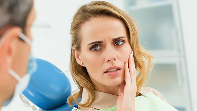 Emergency Dentist A concerned woman with blonde hair is sitting in a dental chair, touching her cheek with a pained expression. She appears to be discussing her discomfort with a dentist, who is partially visible in the foreground wearing a mask. The setting is a dental clinic with medical equipment in the background.