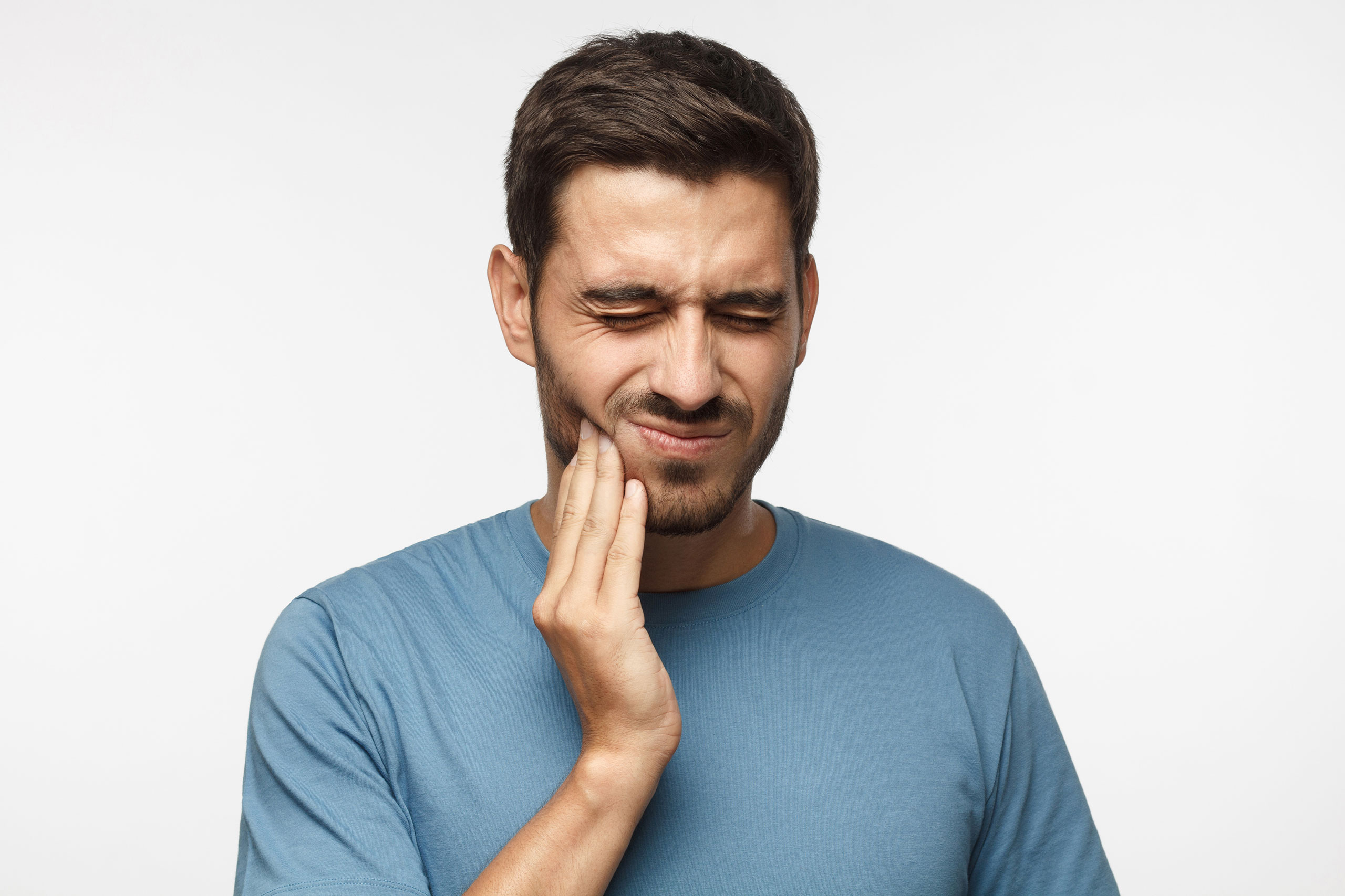 Man in a blue t-shirt with a pained expression, eyes closed and hand pressed against his cheek, indicating gum pain, toothache or jaw discomfort. The background is plain white, keeping focus on the man's facial expression and gesture.