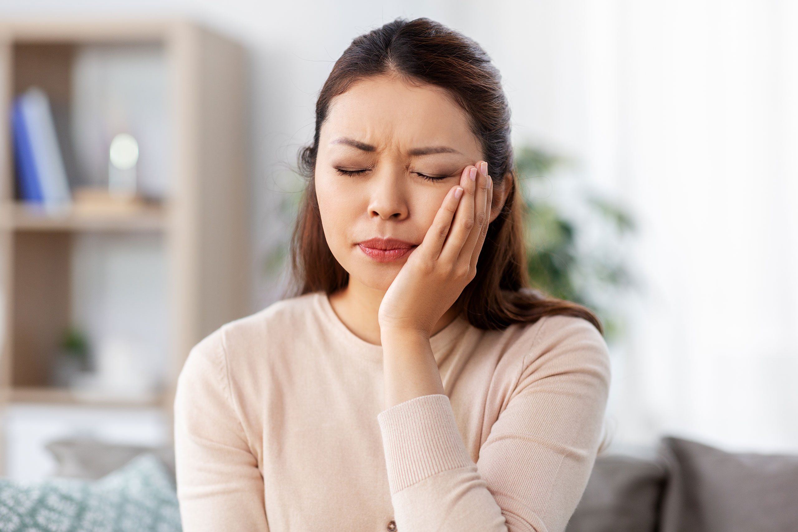 Woman sitting indoors with a pained expression, eyes closed and one hand pressing against her cheek, indicating tooth or jaw pain. She is wearing a light beige top, and the background features a softly focused living room setting.