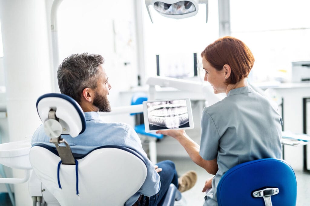 Female dentist showing a male patient a dental X-ray on a tablet while they sit in a bright, modern dental office; both are smiling and engaged in conversation.