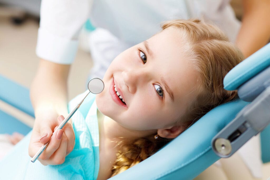 Smiling young girl lying back in a blue dental chair, holding a dental mirror tool while looking up at the camera, with a dental professional partially visible in the background.
