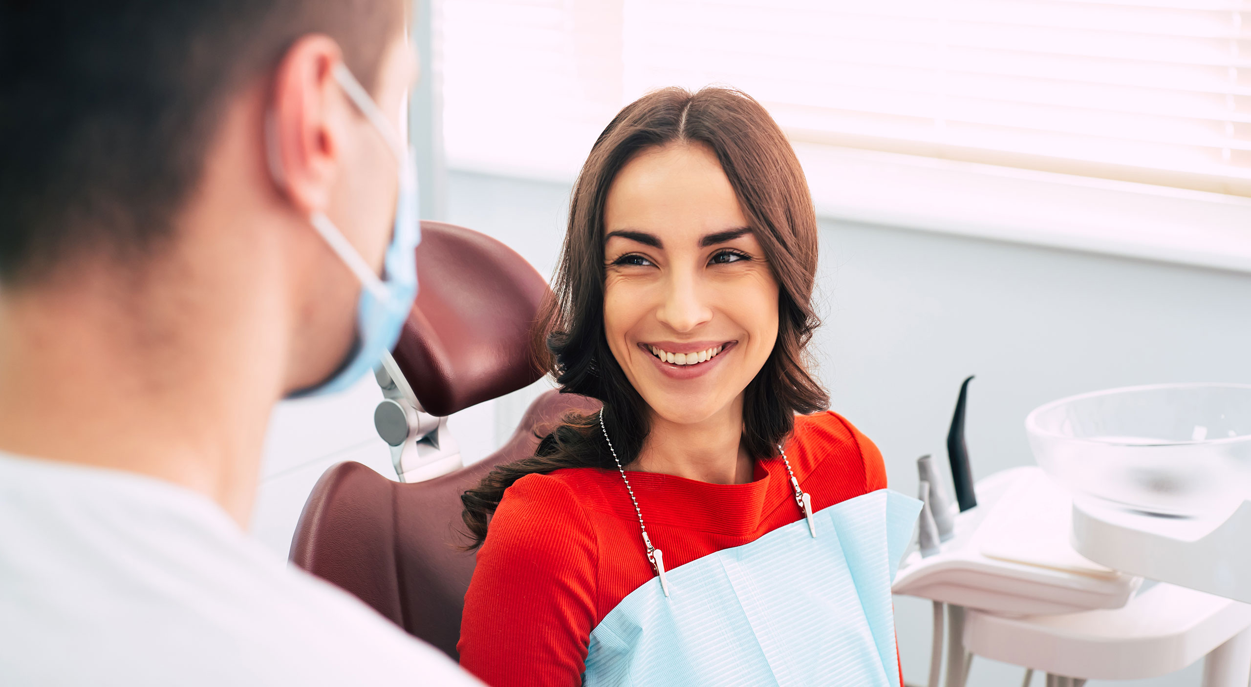 Smiling woman sitting in a dental chair wearing a red top and a blue dental bib, looking at a male dentist who is wearing a surgical mask, inside a brightly lit dental office.