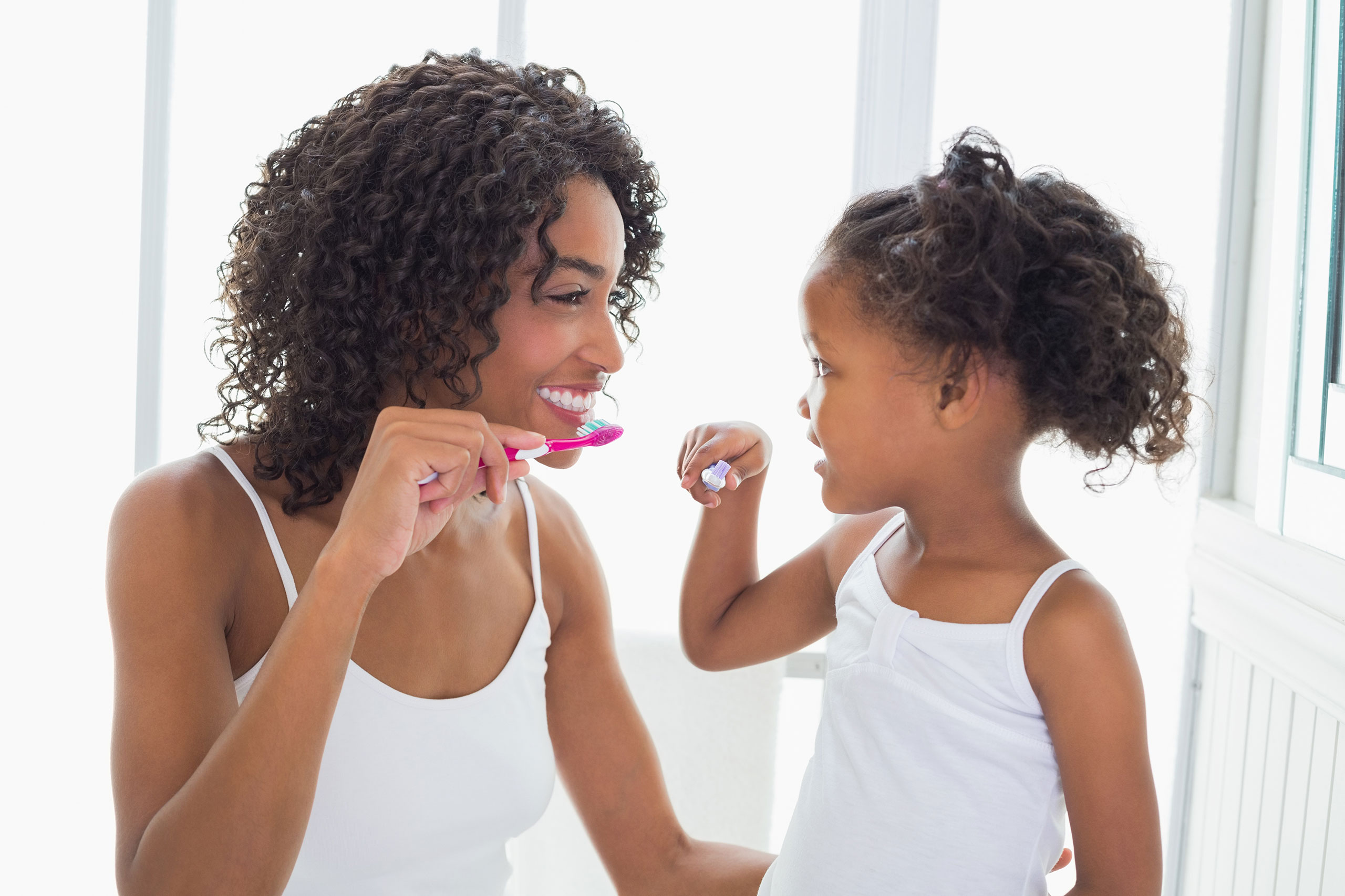 Smiling woman and young girl, both wearing white tank tops, facing each other and holding toothbrushes as they brush their teeth together in a bright bathroom.