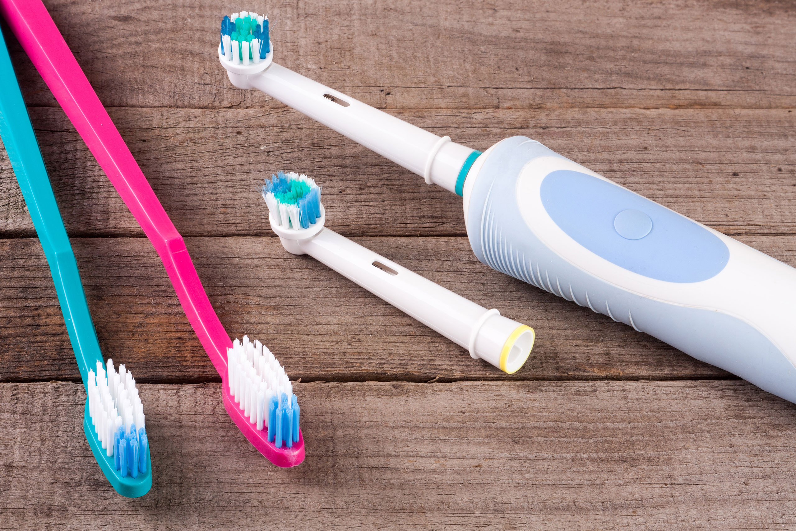 Two manual toothbrushes (one pink and one teal) and an electric toothbrush with two interchangeable brush heads on a rustic wooden surface.