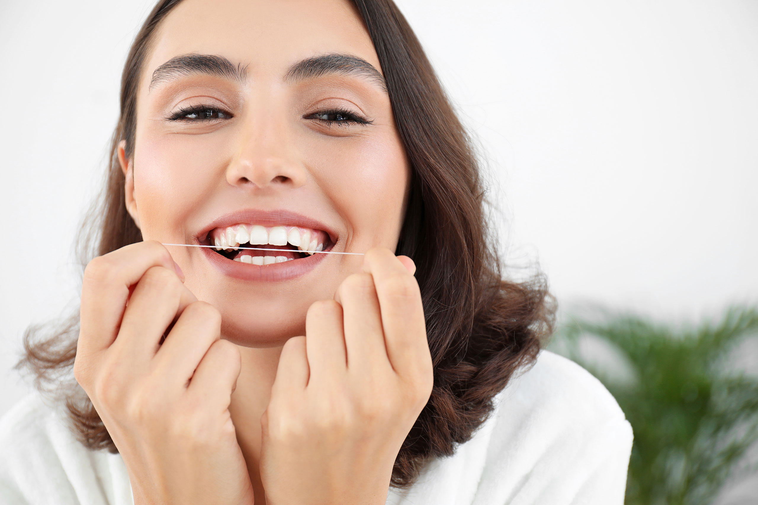 Close-up of a smiling woman with brown hair using dental floss to clean her teeth, wearing a white robe, with a blurred green plant in the background.