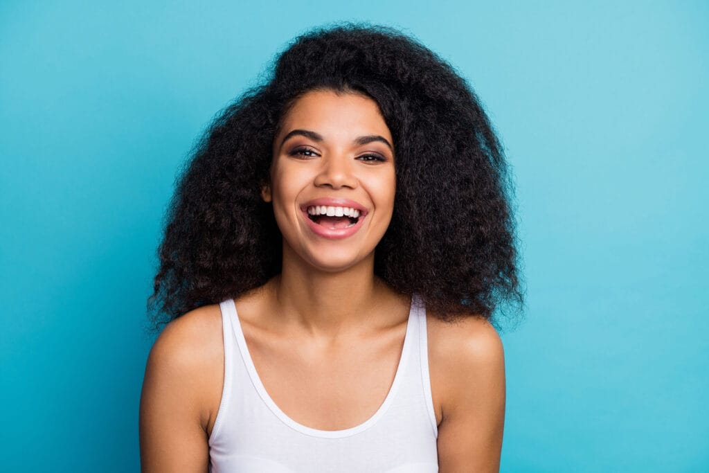 Smiling young woman with voluminous curly hair wearing a white tank top, posing against a bright blue background.