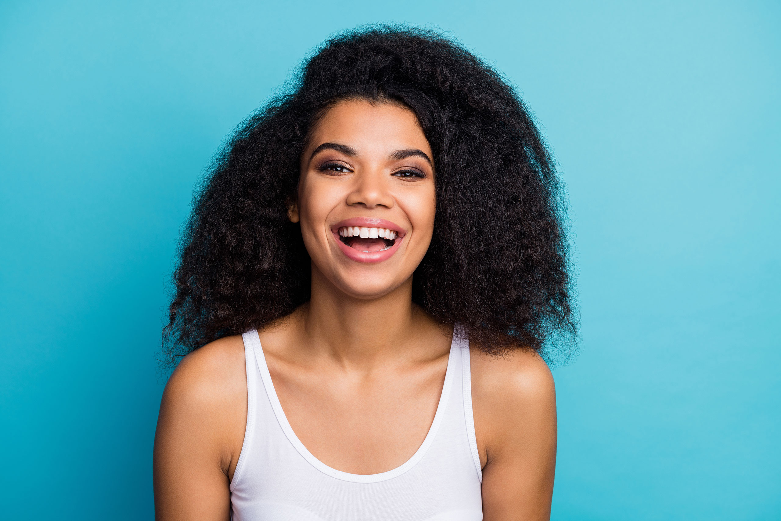 Smiling young woman with voluminous curly hair wearing a white tank top, posing against a bright blue background.