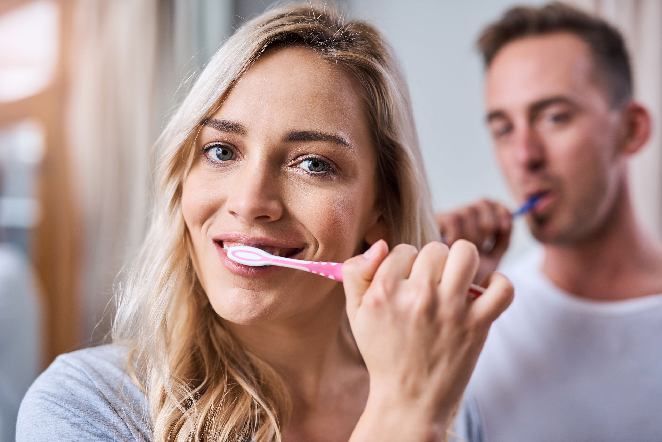 Smiling woman brushing her teeth with a pink toothbrush in the foreground; man blurred in the background brushing with a blue toothbrush in a bright bathroom.