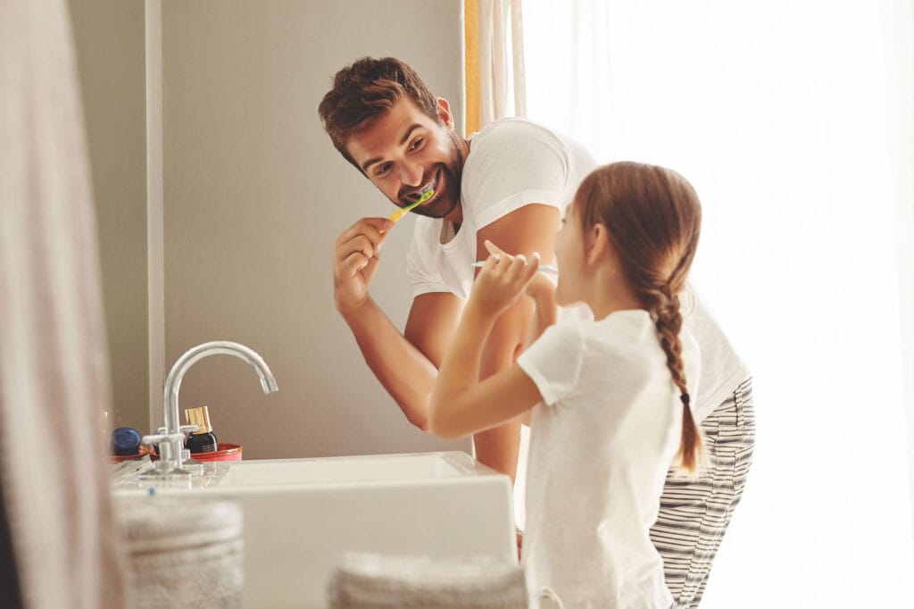 Smiling man and young girl in white shirts brushing their teeth together in a bright bathroom, standing by a sink with a silver faucet and toiletries in the background.