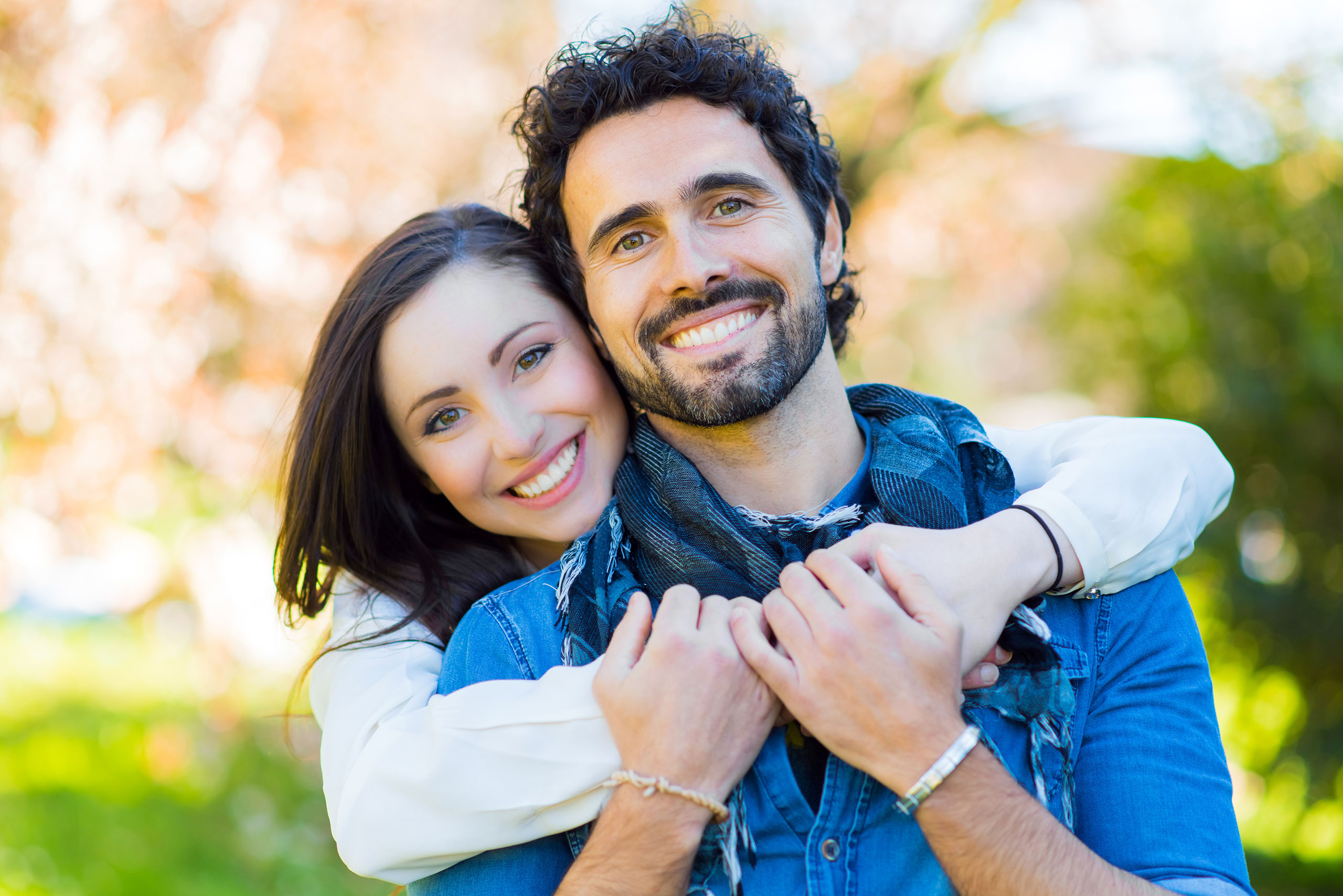 Smiling couple embracing outdoors, with the woman in a white top hugging the man from behind. Both have dark hair, bright smiles, and are dressed casually, standing in front of a blurred background of greenery and sunlight.