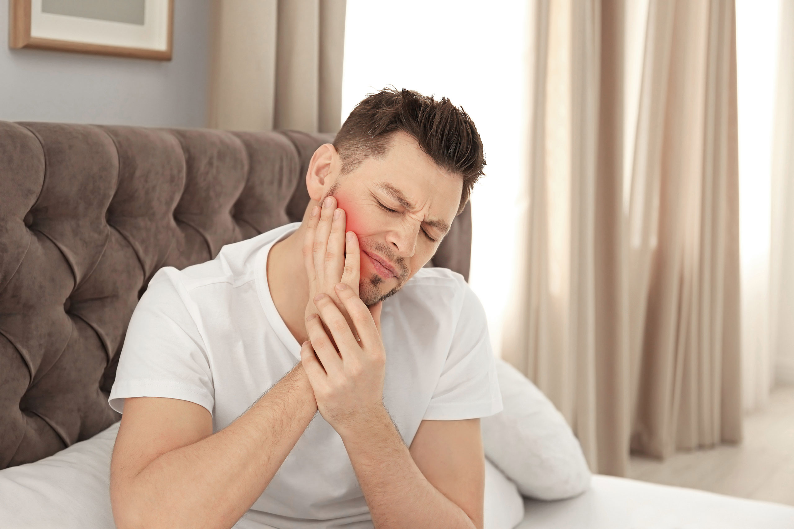 Man sitting on bed holding his cheek with a pained expression, showing discomfort from tooth or jaw pain from teeth grinding.