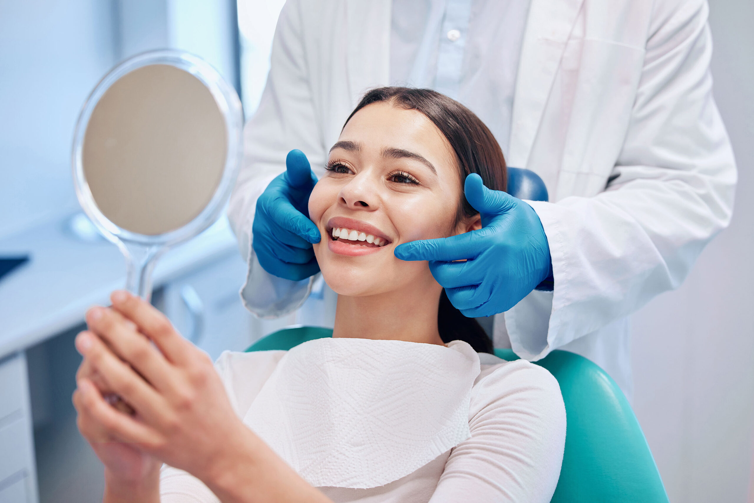 Smiling dental patient holding a mirror while dentist in gloves checks her teeth and gums during consultation.