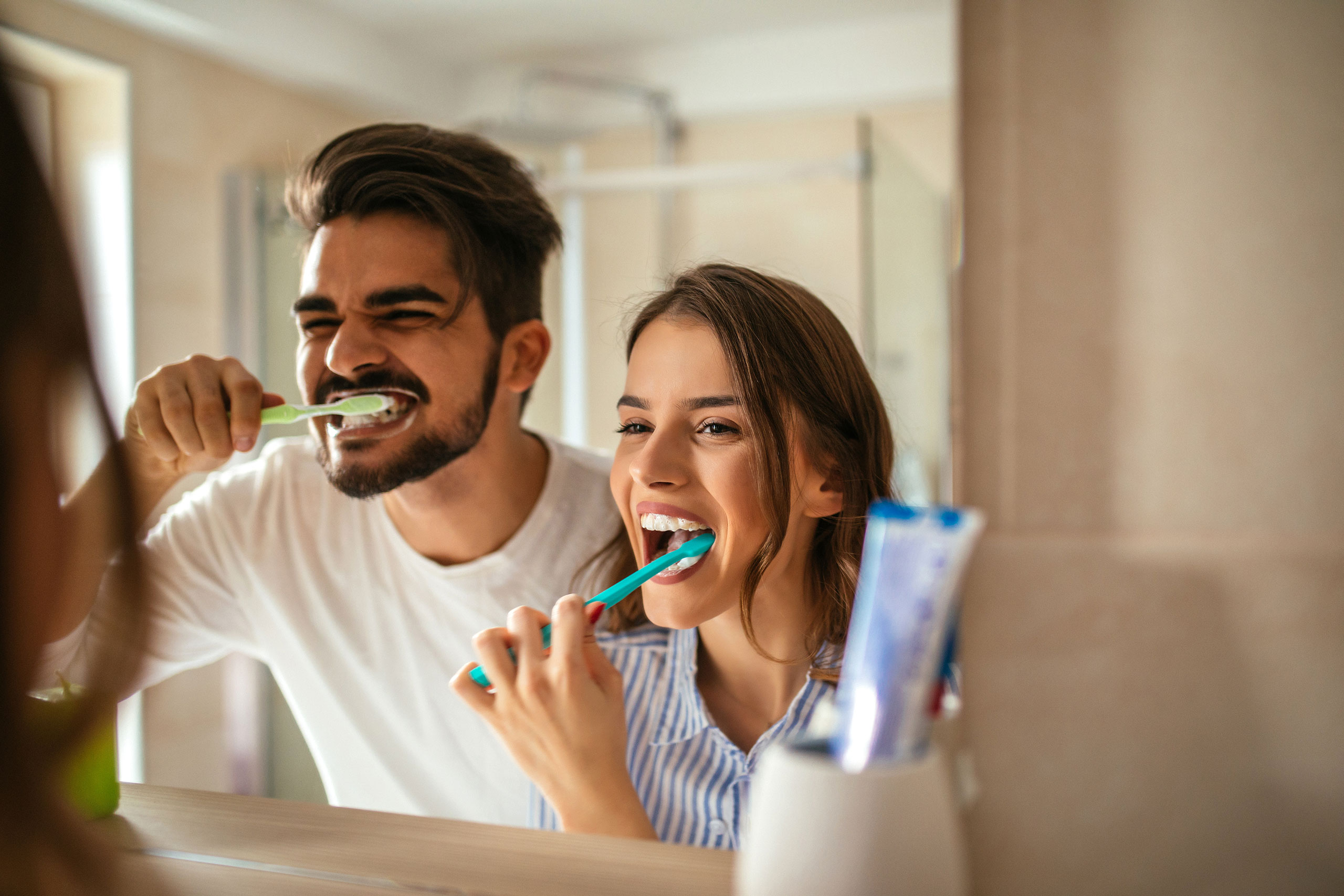 Smiling couple brushing their teeth together in front of a bathroom mirror in the morning.