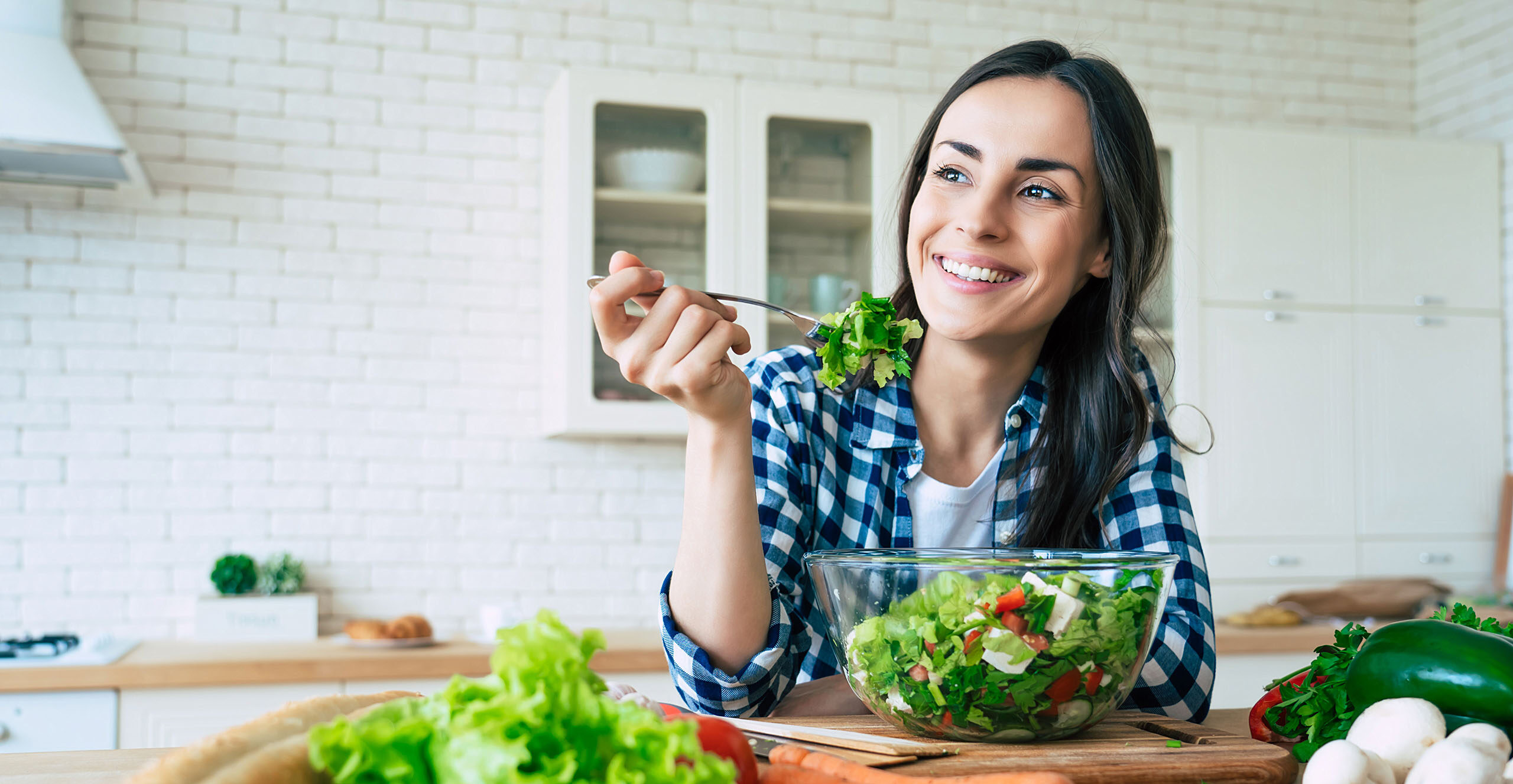 Smiling young woman wearing a blue and white plaid shirt, eating a fresh green salad in a bright kitchen with assorted vegetables on the counter.