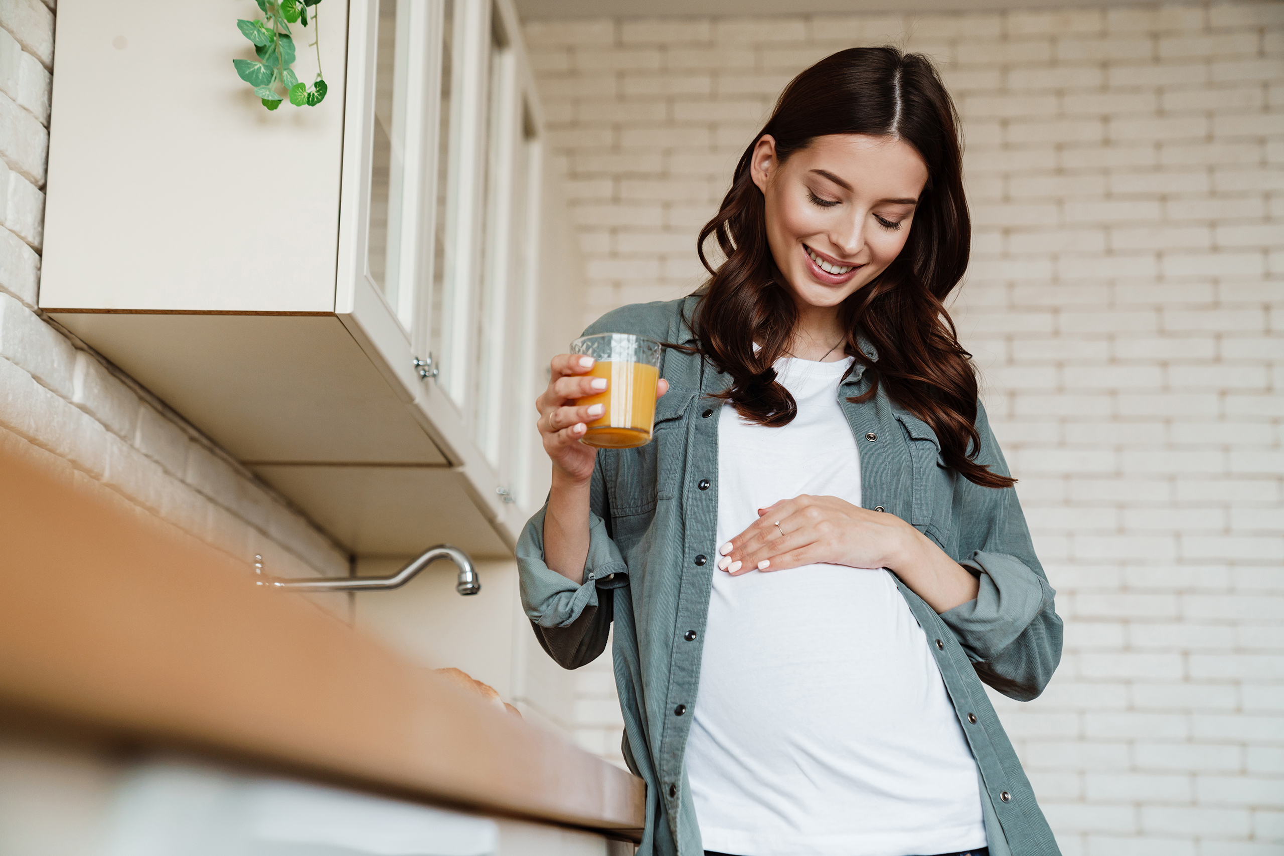 Smiling pregnant person in a kitchen holding a glass of orange juice while gently touching their belly.
