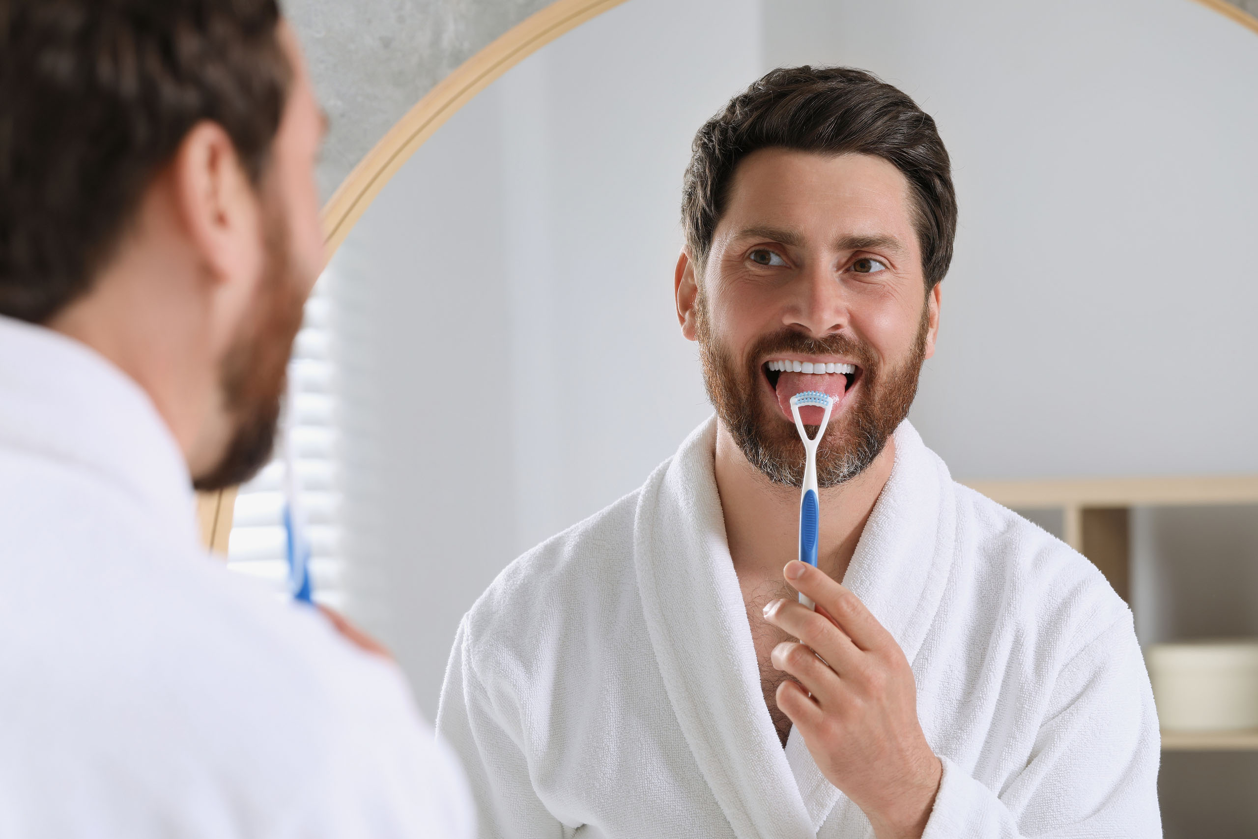 Man in a white bathrobe using a tongue scraper in front of a bathroom mirror as part of his oral hygiene routine.