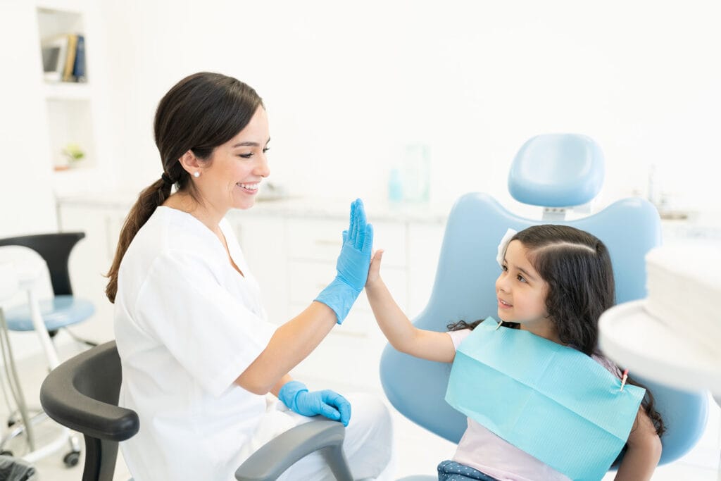 Pediatric dentist wearing gloves gives a high five to a smiling young patient seated in a dental chair during a friendly visit.