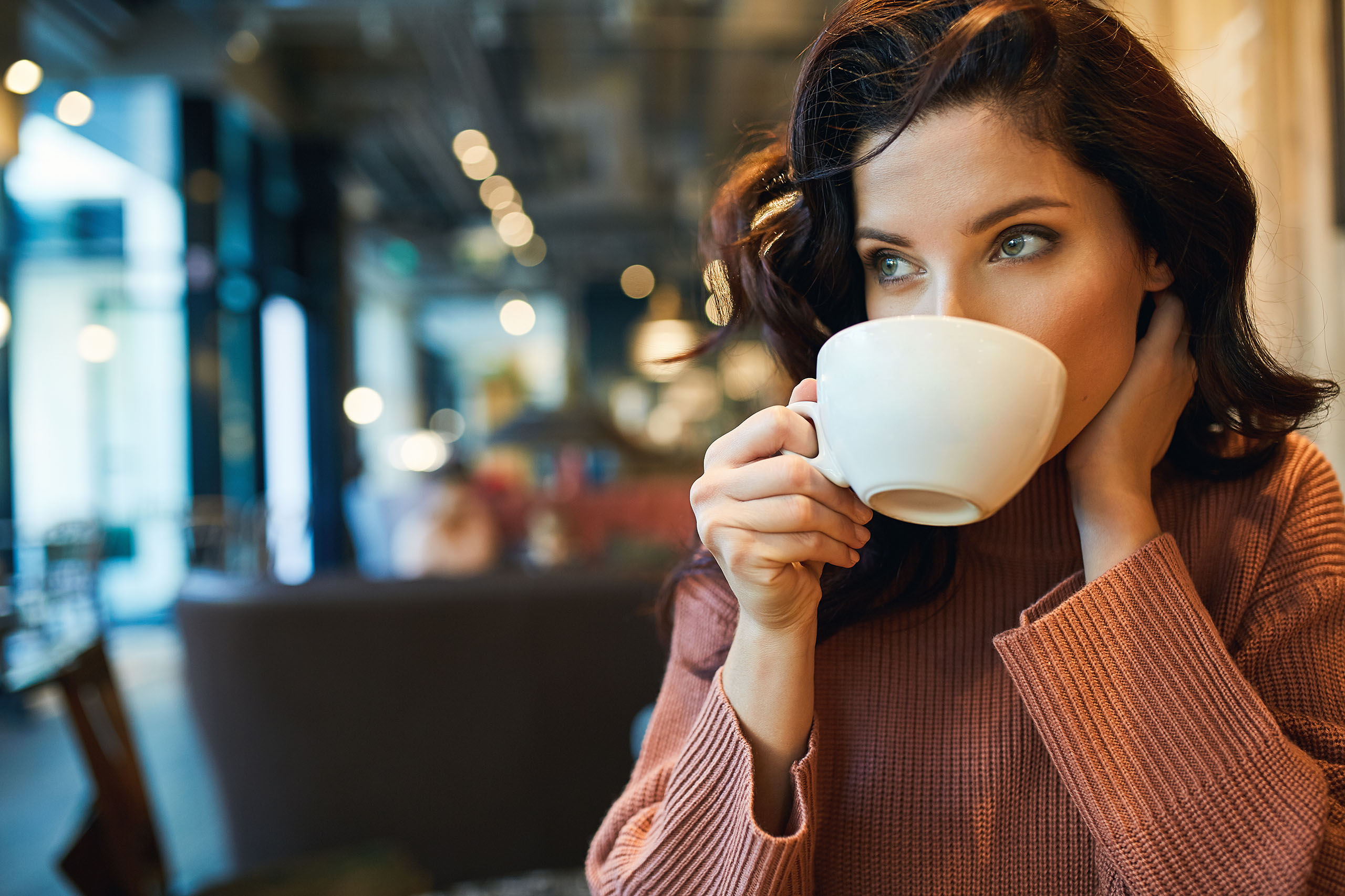 Woman sipping a hot drink in a café, holding a cup near her mouth with a thoughtful expression