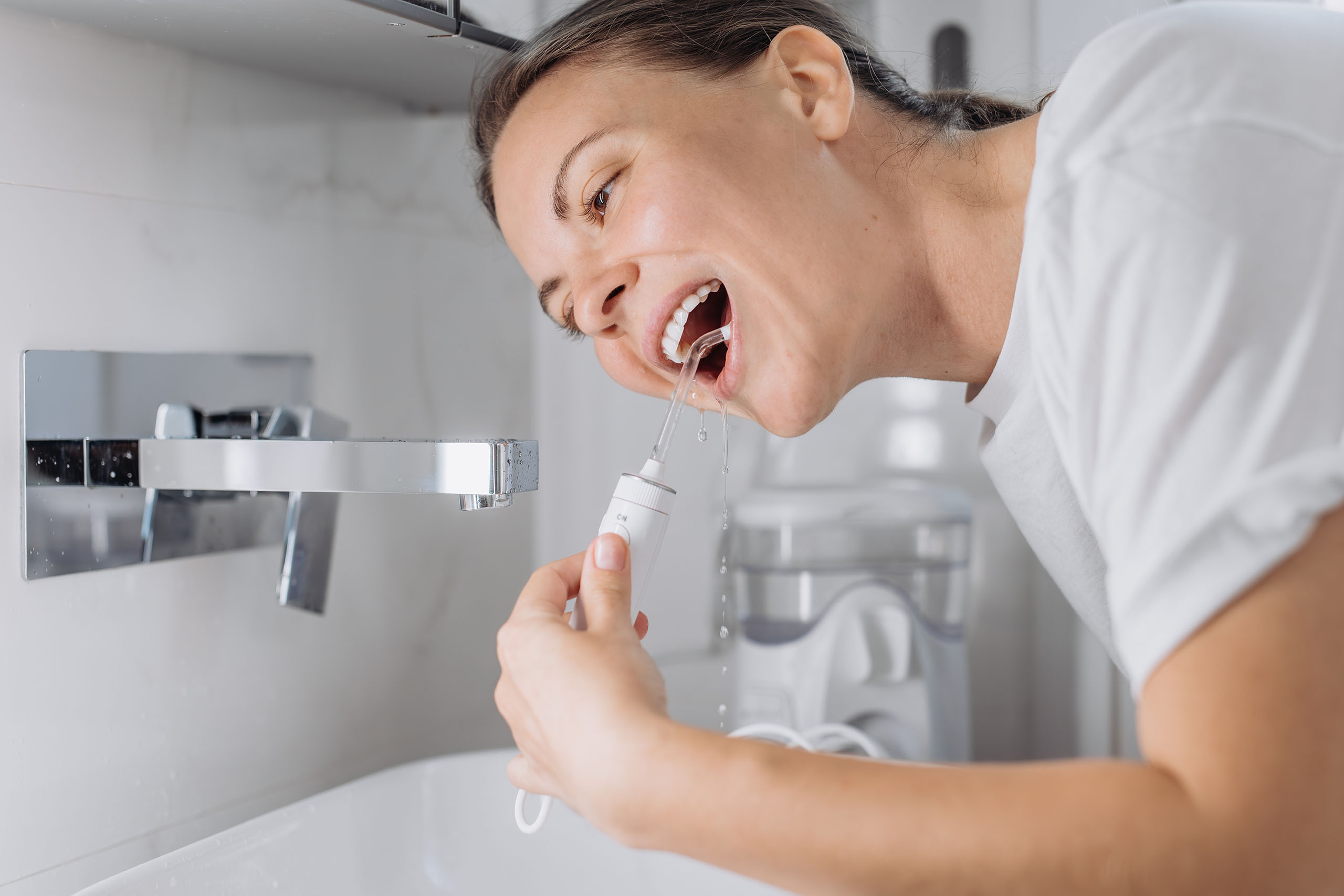 Person using a water flosser at a bathroom sink, directing a stream of water between teeth for oral hygiene