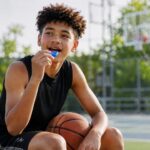 Teen athlete seated on an outdoor basketball court in bright sunlight, holding a basketball while lifting a blue custom mouthguard toward his mouth, with an open mouthguard case beside him.