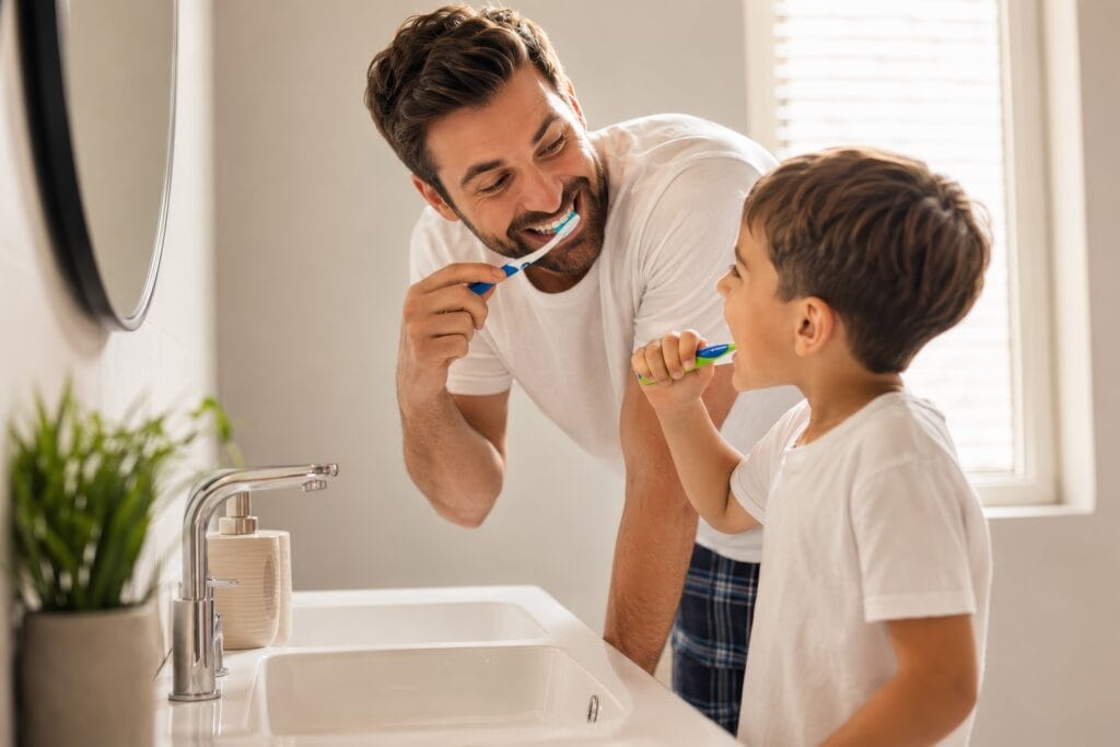 Father and young child brushing their teeth together in a bright, modern bathroom, smiling at each other beside a white sink in warm natural light.